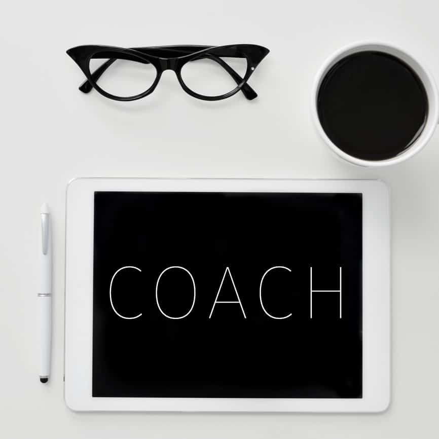 Black glasses, coffee cup and ipad with the word "Coach" on it, symbolizing "Coaching."