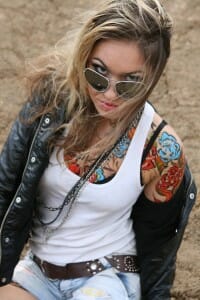 Young woman in a tank top, blue jean shorts, and a leather jacket showing a shoulder and chest full of tattoos.