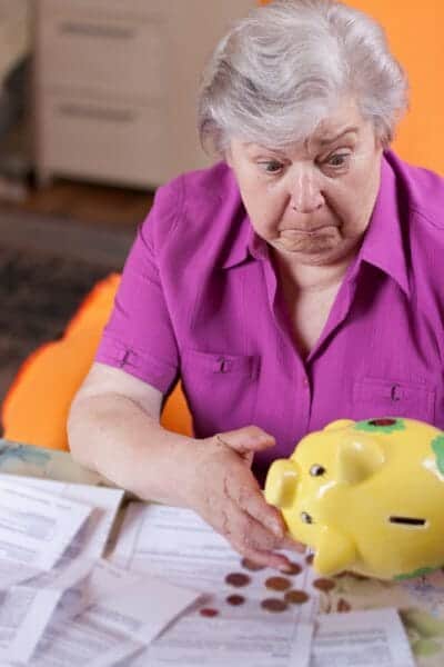 Worried senior woman shaking pennies out of a yellow piggy bank onto the paperwork on her desk.