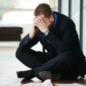 Business man sitting on the floor of his office with his head in his hands.