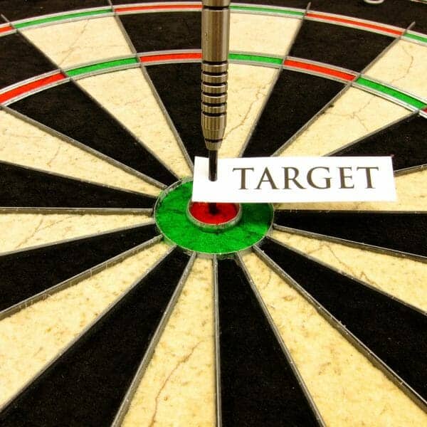 Close up of a dart board with a dart standing up in the bull's eye with a note that says "Target" on it.