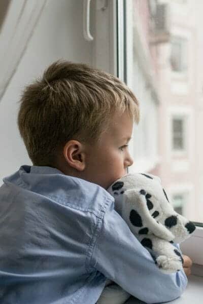 Sad boy holding a stuffed toy and looking out the window.