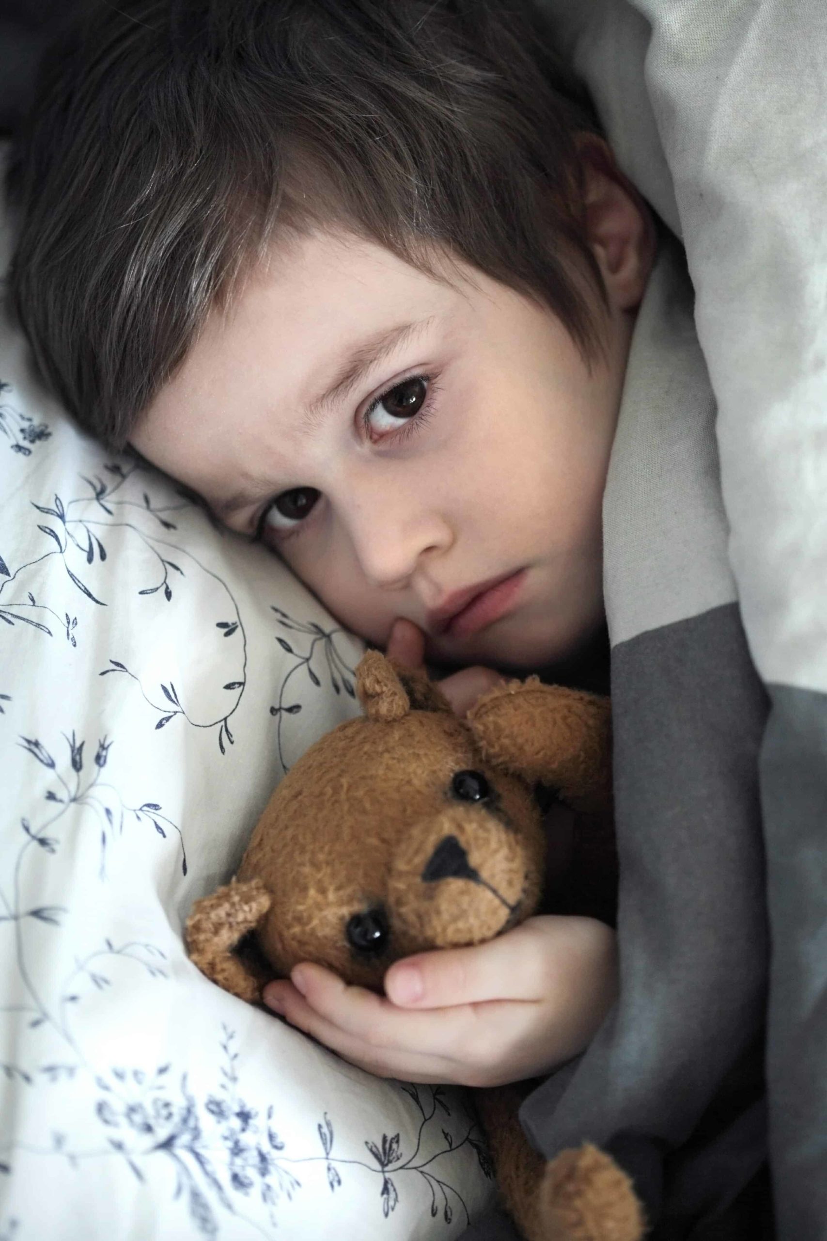 Small sad boy in bed, holding his teddy bear.