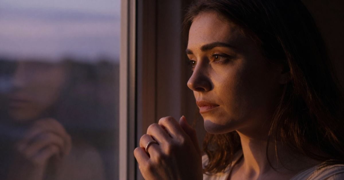 AI image of a woman looking out a window at dusk, looking solemn and conflicted