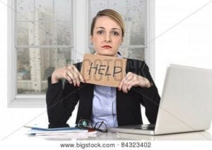 Sad business woman holding a "Help" sign as she sits at her computer.