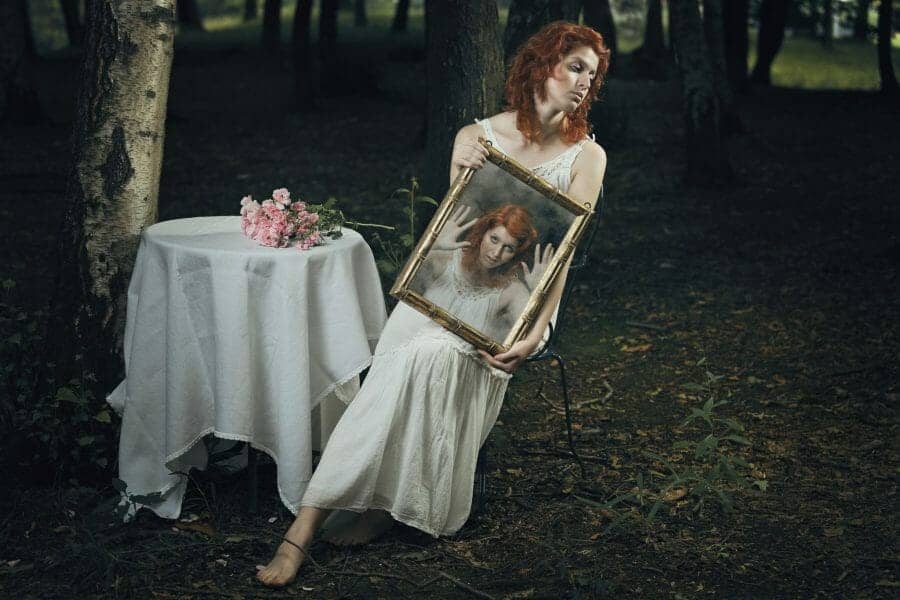 Young woman in a white dress in the forest holds a picture of herself trying to get out of the picture frame