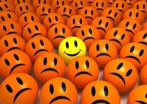 One yellow happy smiling emoticon in the middle of a crowd of orange sad emoticons