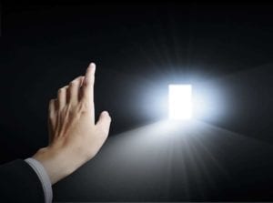 Black room with a light streaming in from a far door with a hand pointing to the door.