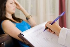 Close up of a therapists hand taking notes while a troubled woman rests on a couch in front of her. Counseling.