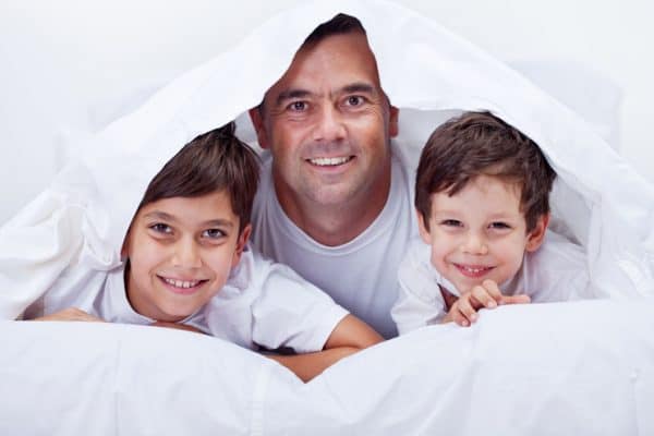 Smiling picture of Dad and his boys hiding under the covers in a white bed