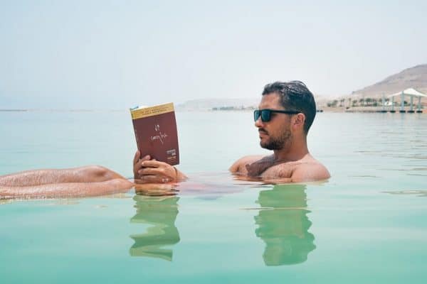 Handsome man with sunglasses floating in tropical water reading a book.