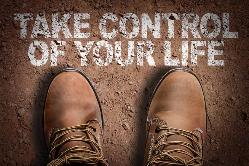 Man's Brown shoes on brown dirt road with the words "TAKE CONTROL OF YOUR LIFE"  on the ground.