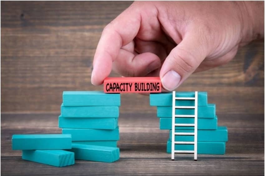 Hand building a small building with blue erasers, capped with a red eraser labeled "capacity building." Signifying building your emotional capacity.