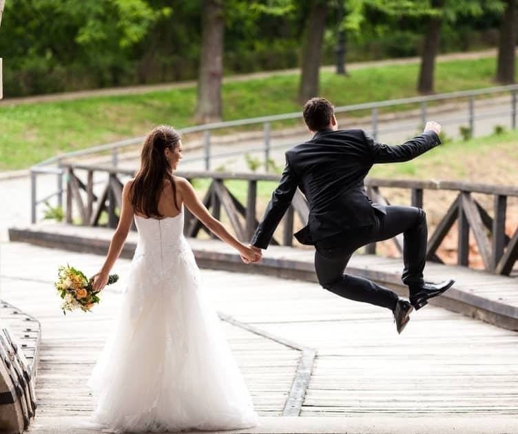 Groom in a second marriage clicking his heels with joy while holding bride's hand and walking down a wooded ramp.