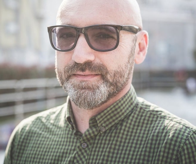 Middle aged bald man with a beard and dark glasses.