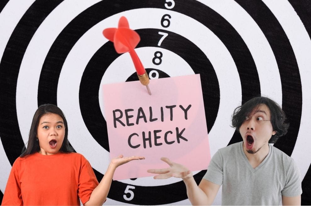 The words "reality check" written on a post it and pinned to a dartboard. A woman and man have surprised facial expressions, gesturing to the reality check